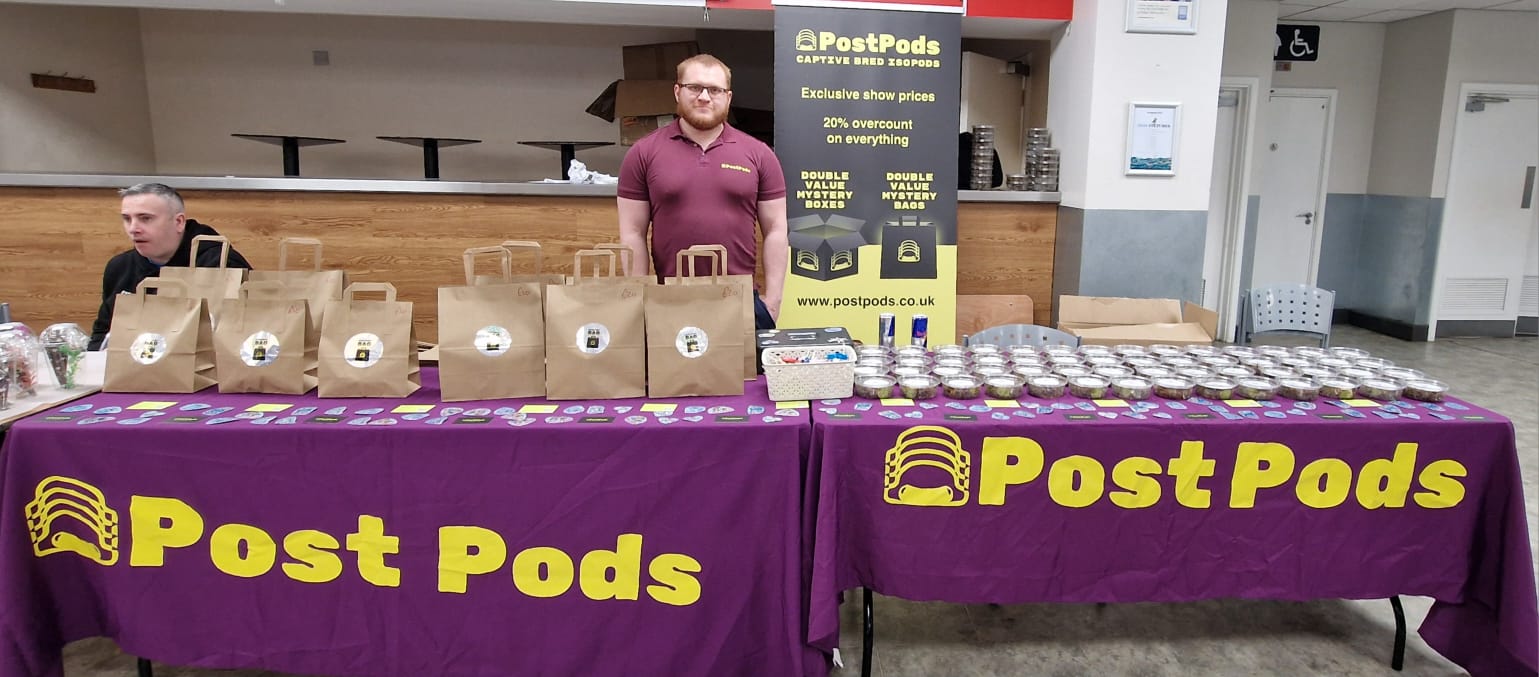 A show stand draped in purple cloths printed Post Pods in yellow, with paper bags, rows of small tubs and two men behind.