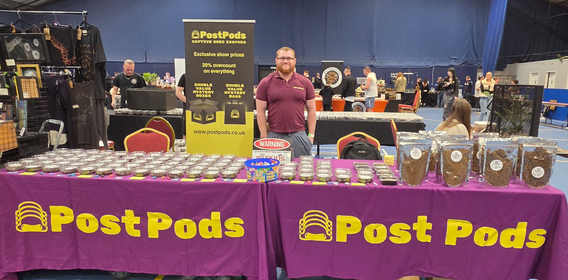 Daniel standing behind the PostPods table at the Coventry Invertebrate Show, with rows of small clear tubs, bags of dry goods and purple PostPods tablecloths