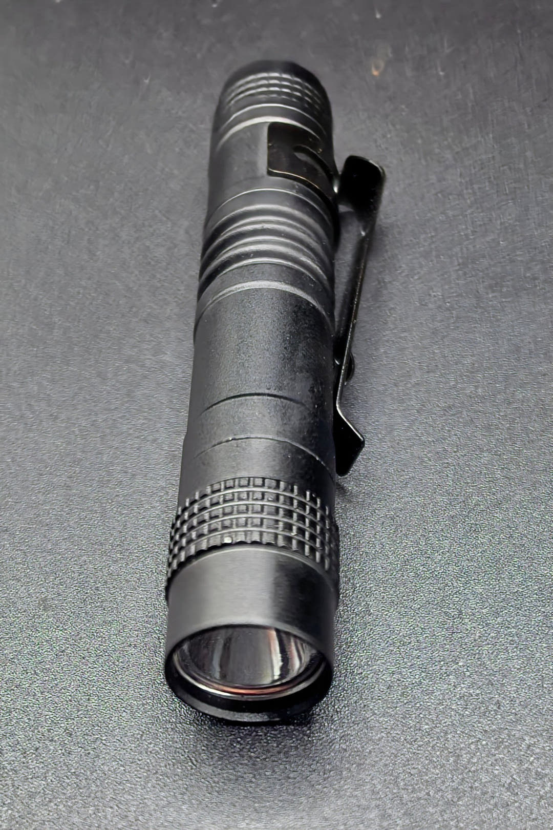 A black tactical flashlight with a textured grip and a black pocket clip, shown on a dark grey textured surface.