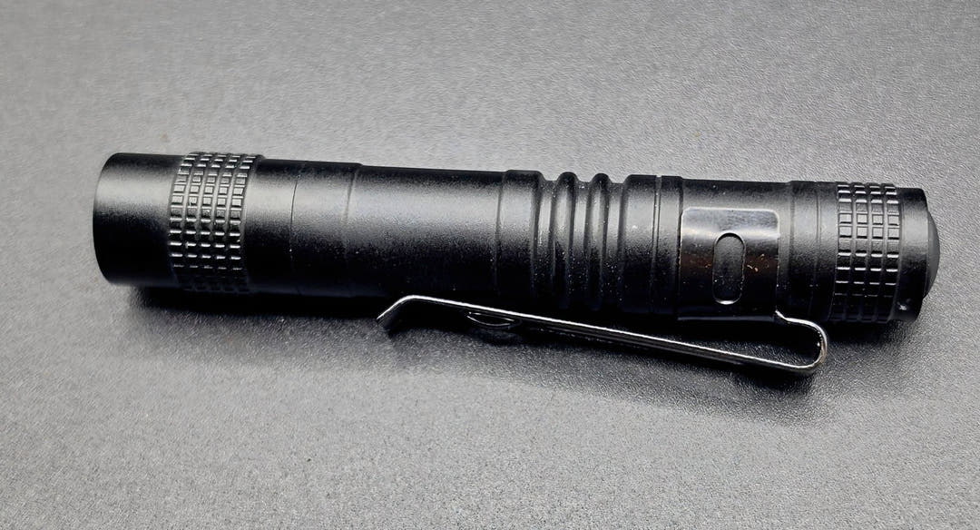 A black tactical flashlight with a built-in metal clip, shown horizontally on a textured grey background.
