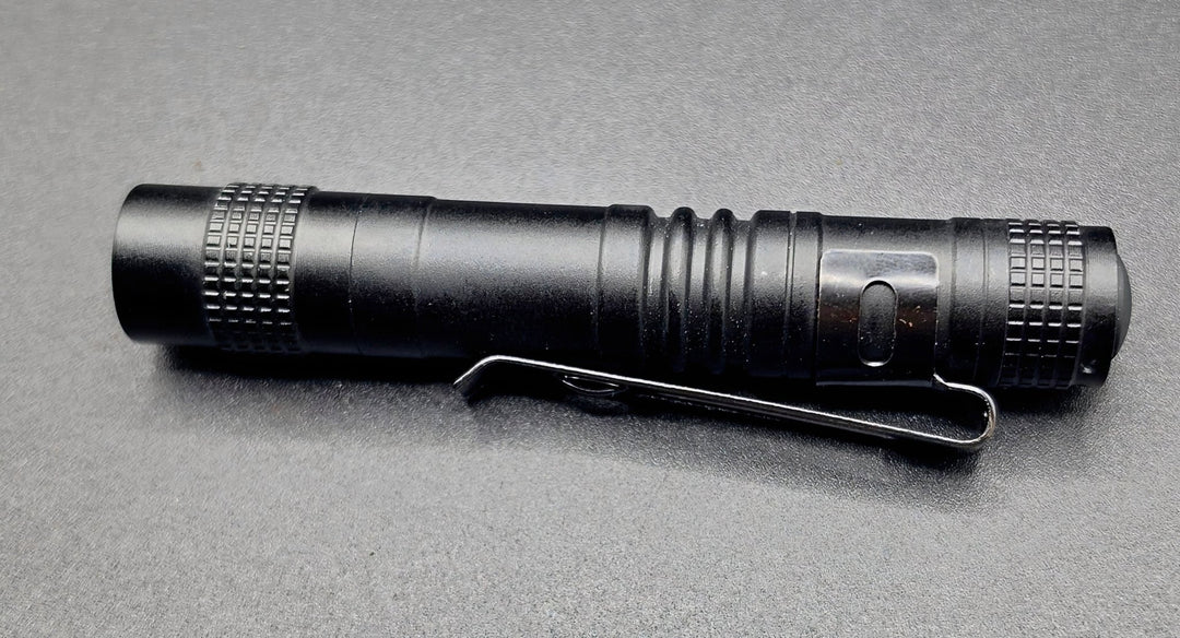 A black tactical flashlight with a built-in metal clip, shown horizontally on a textured gray background.