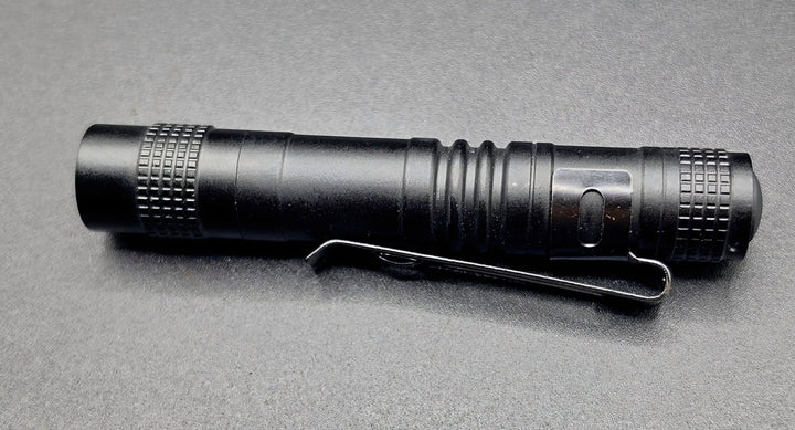 A black tactical flashlight with a built-in metal clip, shown horizontally on a textured gray background.