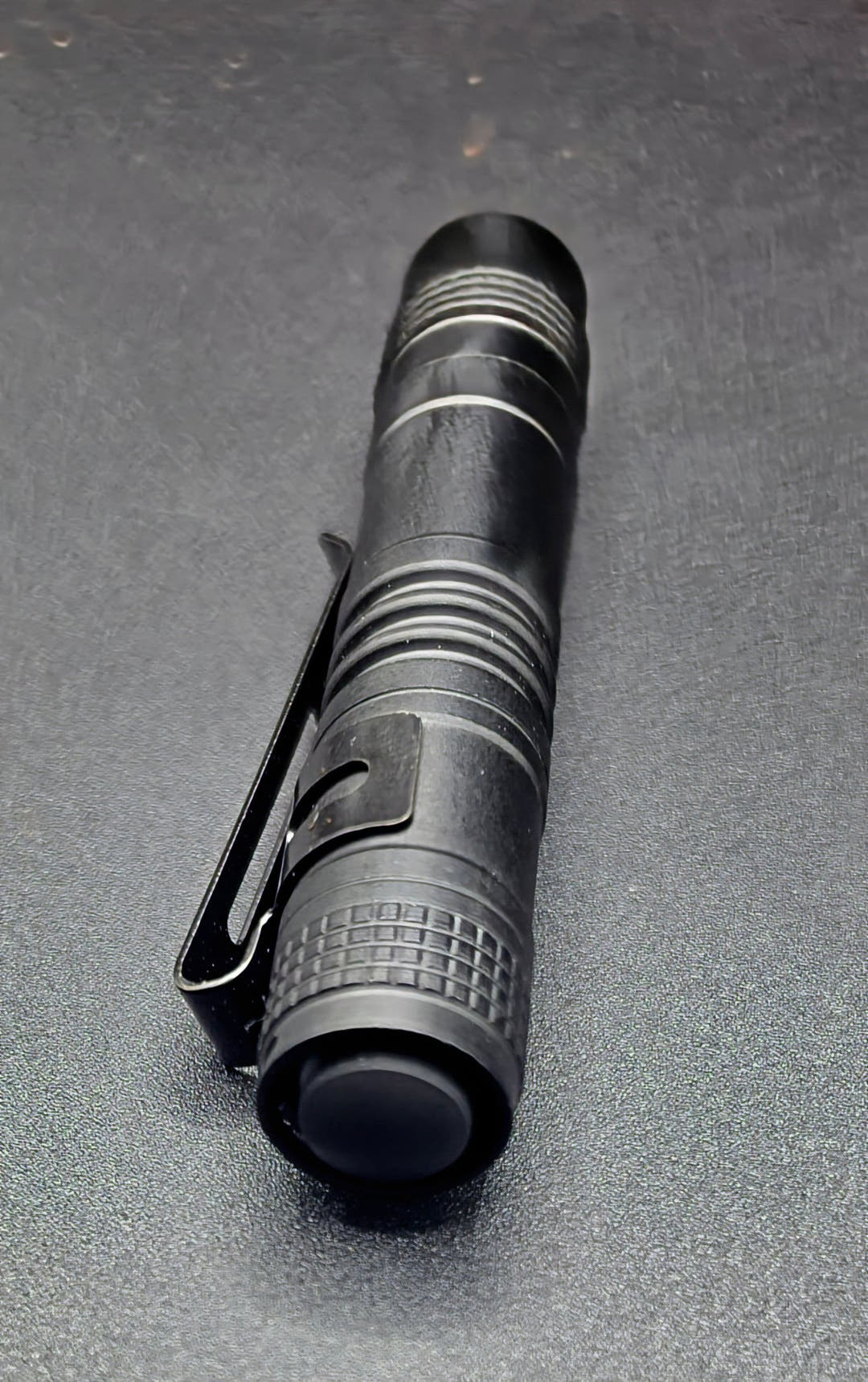 A black tactical pen-style flashlight with a clip, shown diagonally on a dark textured surface.
