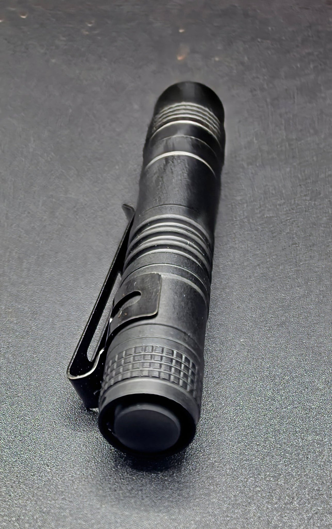 A black tactical pen-style flashlight with a clip, shown diagonally on a dark textured surface.