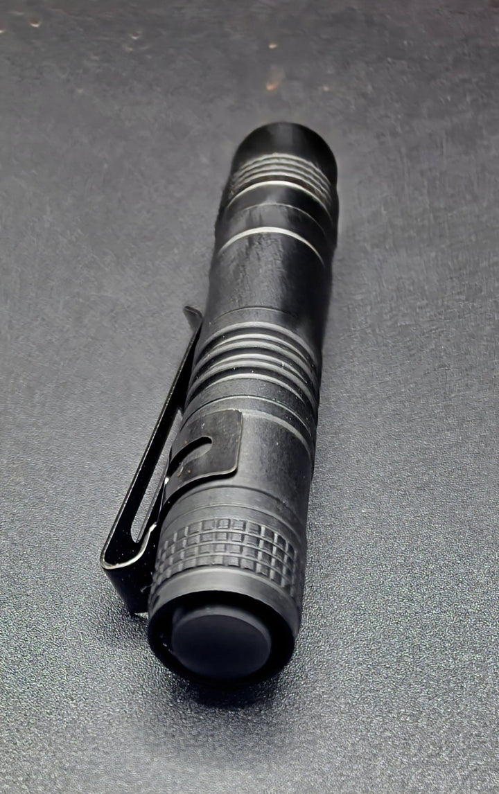 A black tactical pen-style flashlight with a clip, shown diagonally on a dark textured surface.