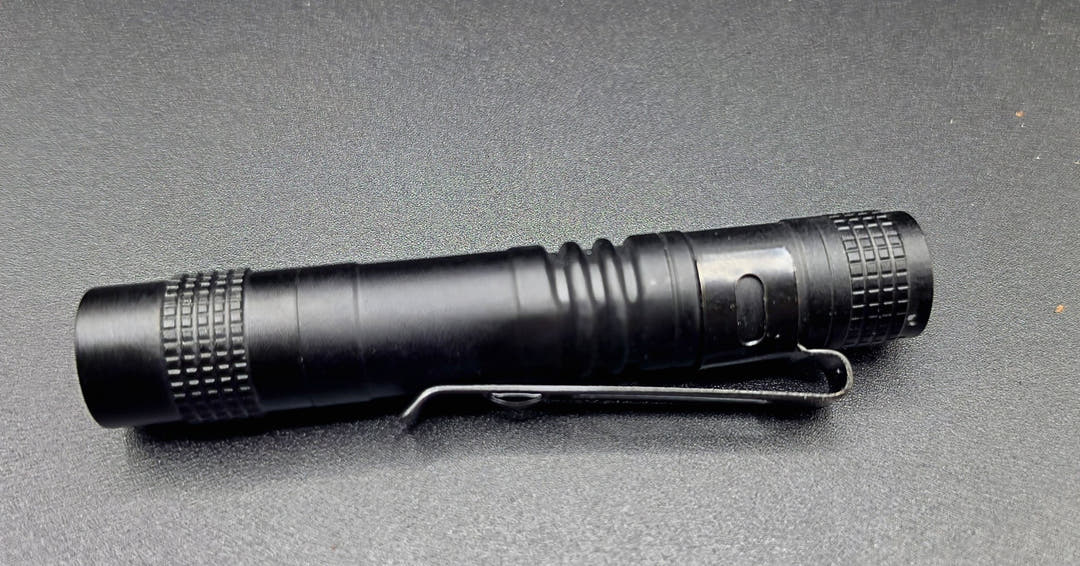 A black tactical flashlight with a built-in pocket clip, resting horizontally on a dark grey textured surface.