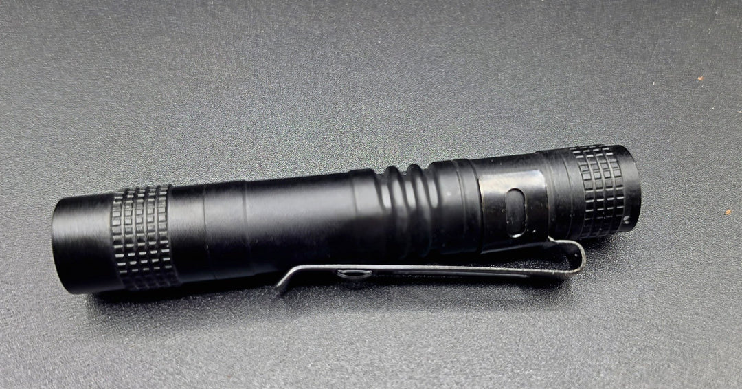 A black tactical flashlight with a built-in pocket clip, resting horizontally on a dark gray textured surface.