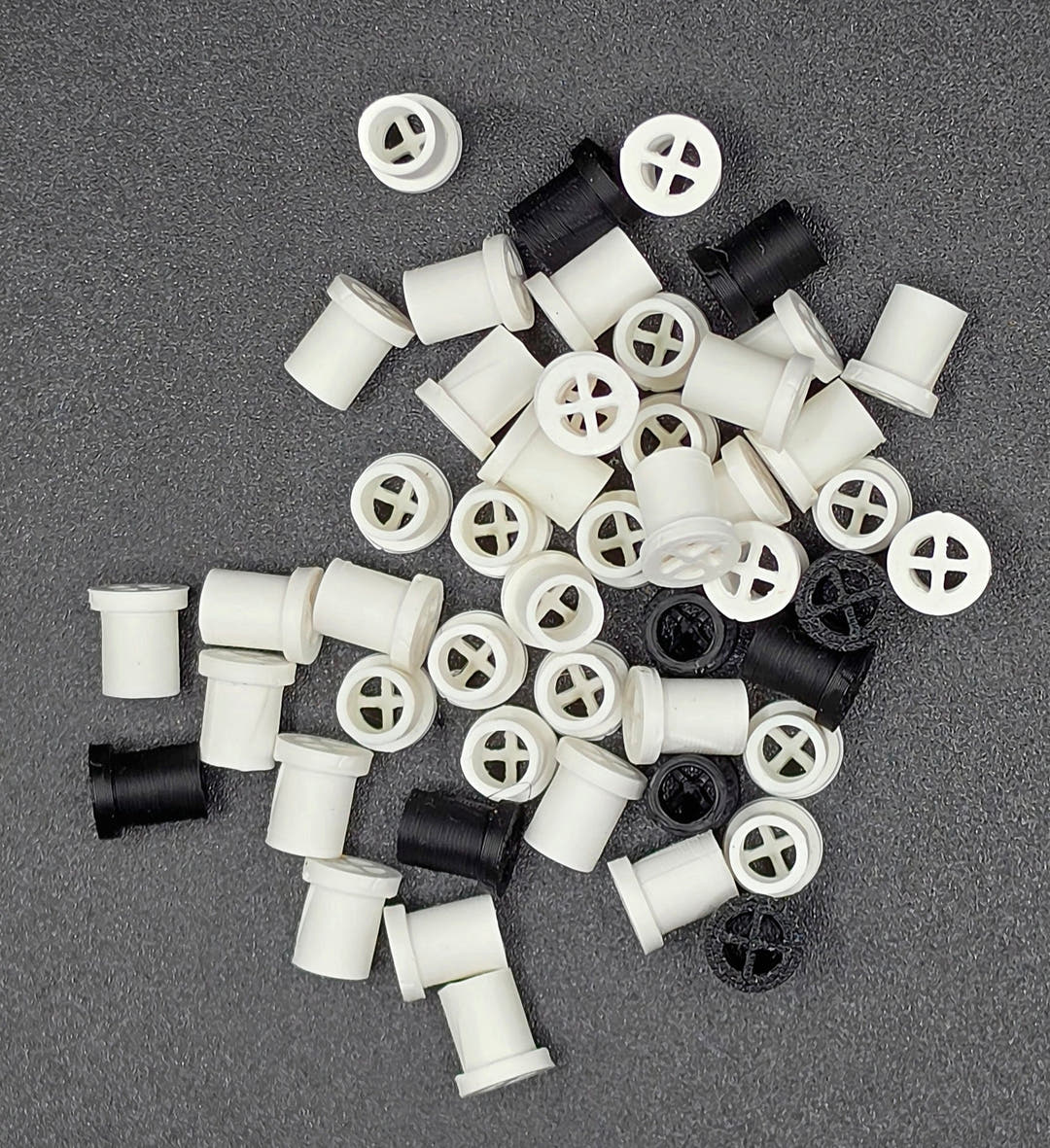 Many white vent plugs with a few black ones spread on a dark grey surface, cross slots across their faces.