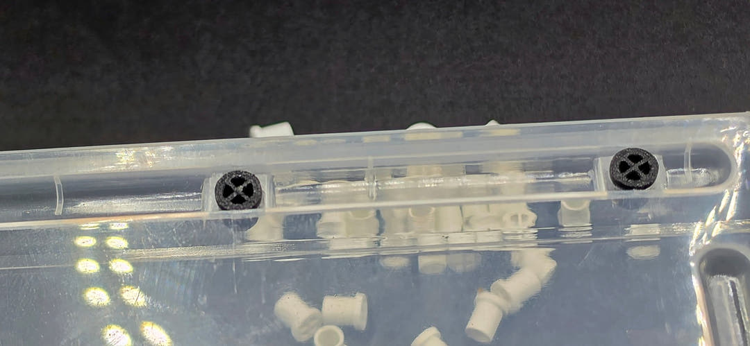 Two black vent plugs fitted through the clear wall of a plastic tub, with pale plugs loose behind it.