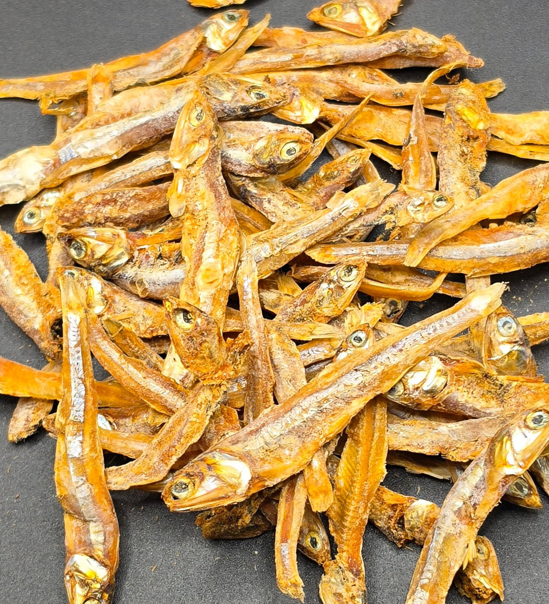 A pile of dried small fish, anchovies, with visible eyes and silvery-brown bodies, spread on a dark gray surface.