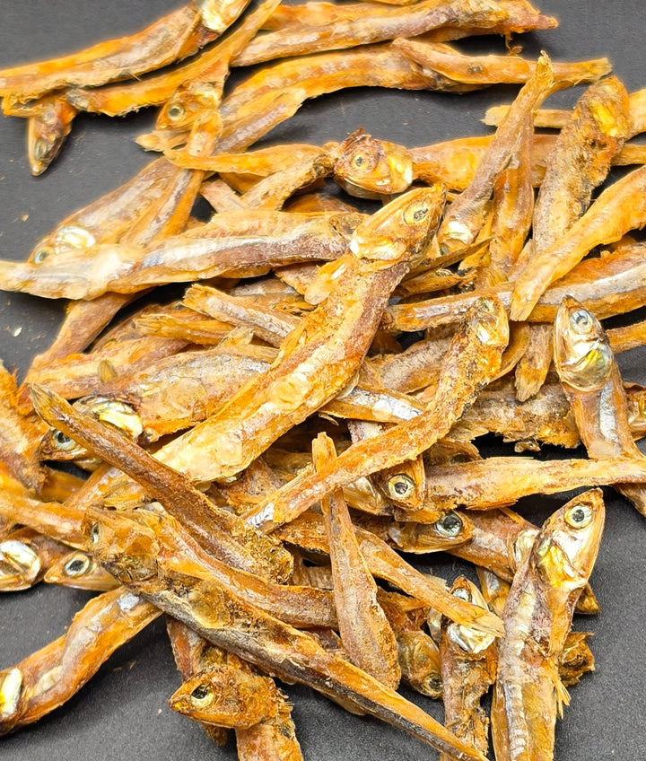 A pile of dried small fish, with visible eyes and golden-brown bodies, arranged on a dark gray surface.