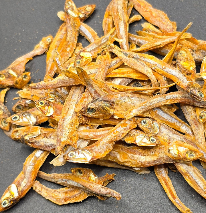 A pile of dried small fish, with visible eyes and golden-brown bodies, arranged on a dark gray background.