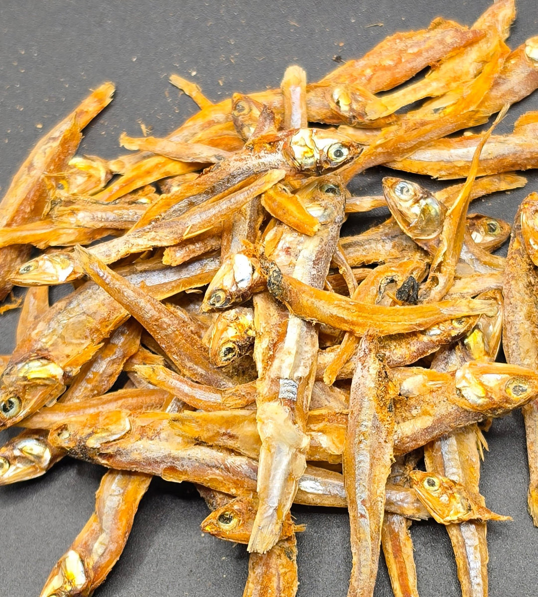 A pile of dried anchovies with visible heads and eyes, spread out on a dark gray surface