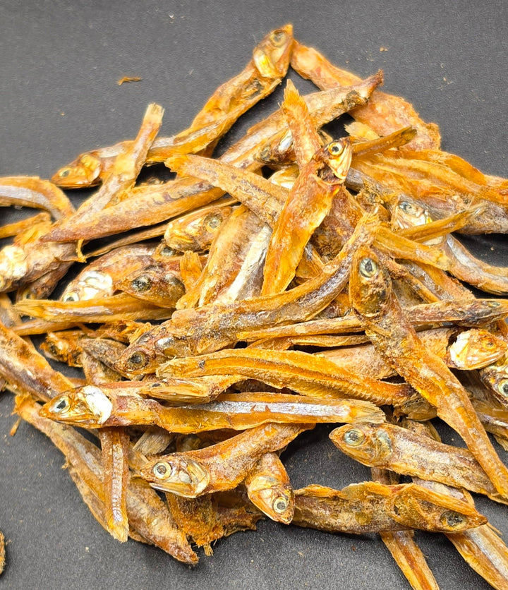 A pile of dried small fish, anchovies, with visible eyes and golden-brown bodies, arranged on a dark gray background.
