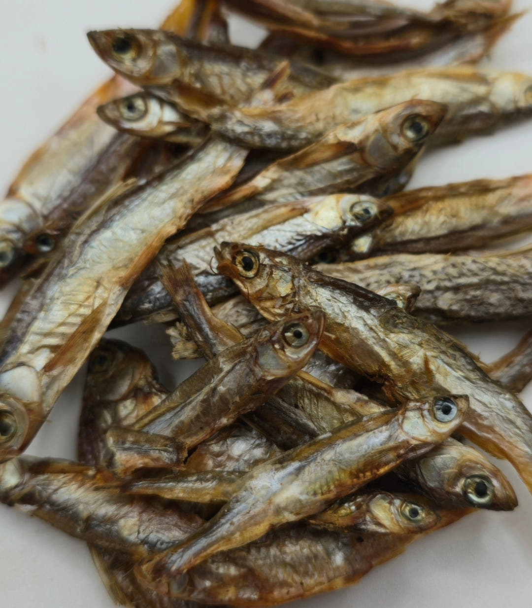 Dried sprats stacked in a mound on white, scales catching the light and eyes clearly visible.