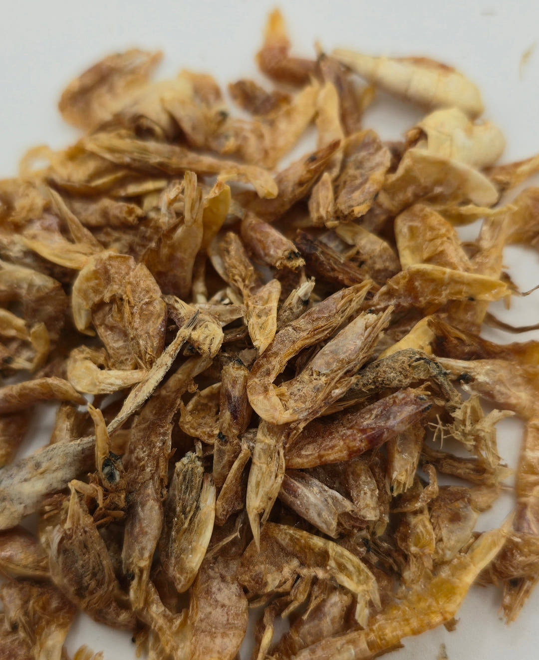 Dried shrimp piled on a white surface, thin golden-brown shells with many broken into pieces.