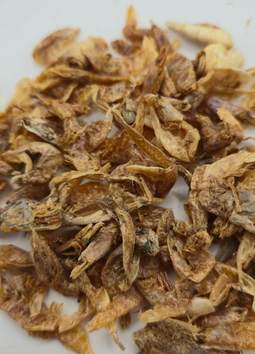 A heap of small dried shrimp in pale gold and brown on a white surface, mostly broken shells and loose heads.