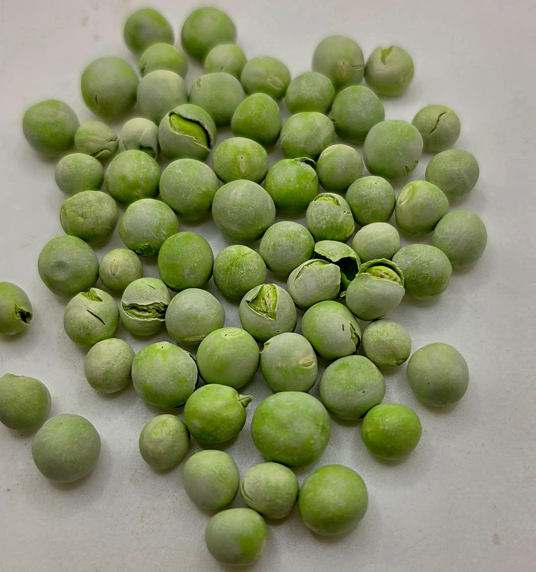 Freeze-dried garden peas spread across a pale surface, a few cracked open to show brighter green inside.