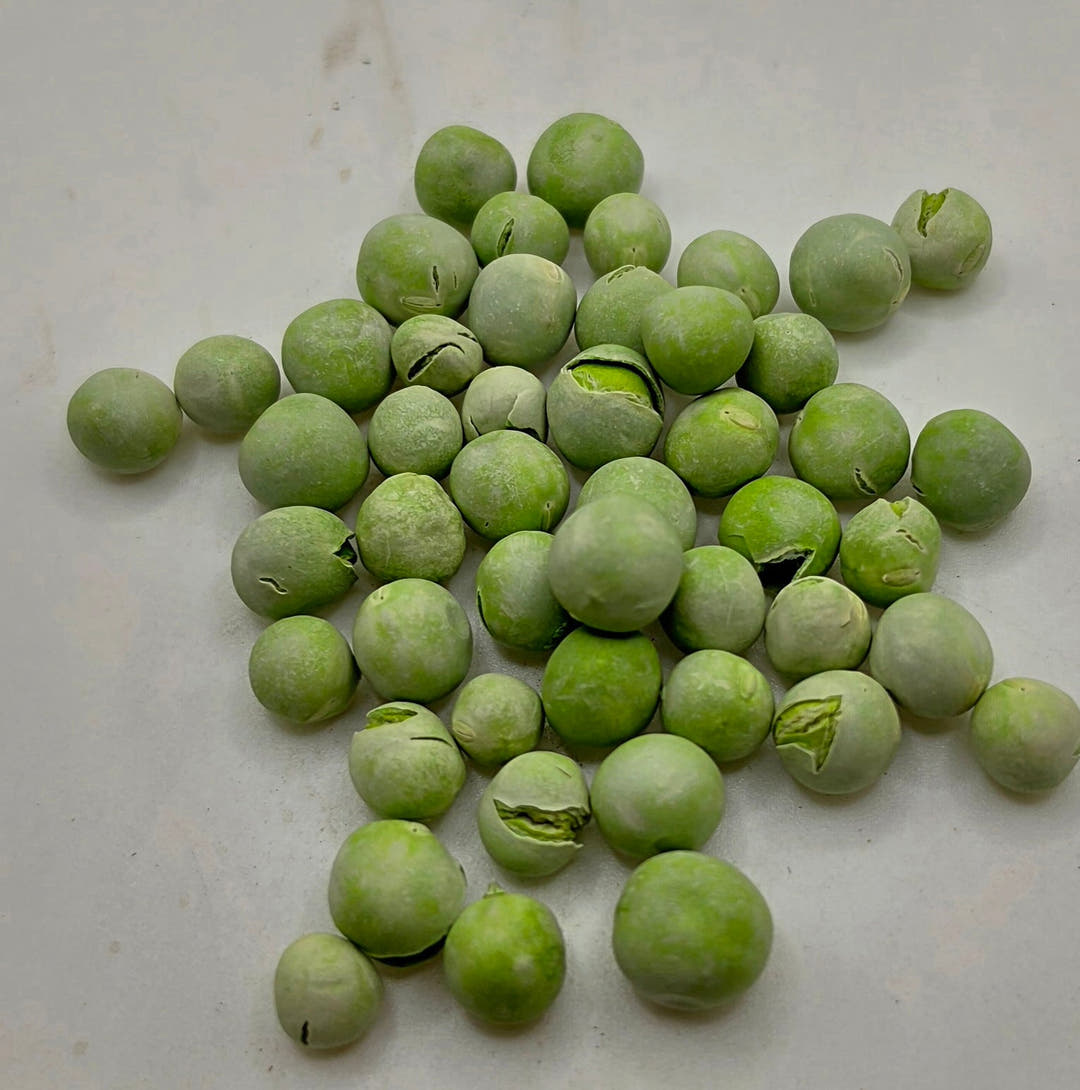Freeze-dried garden peas in a loose scattered pile on a pale grey surface, pale green and dusty looking.