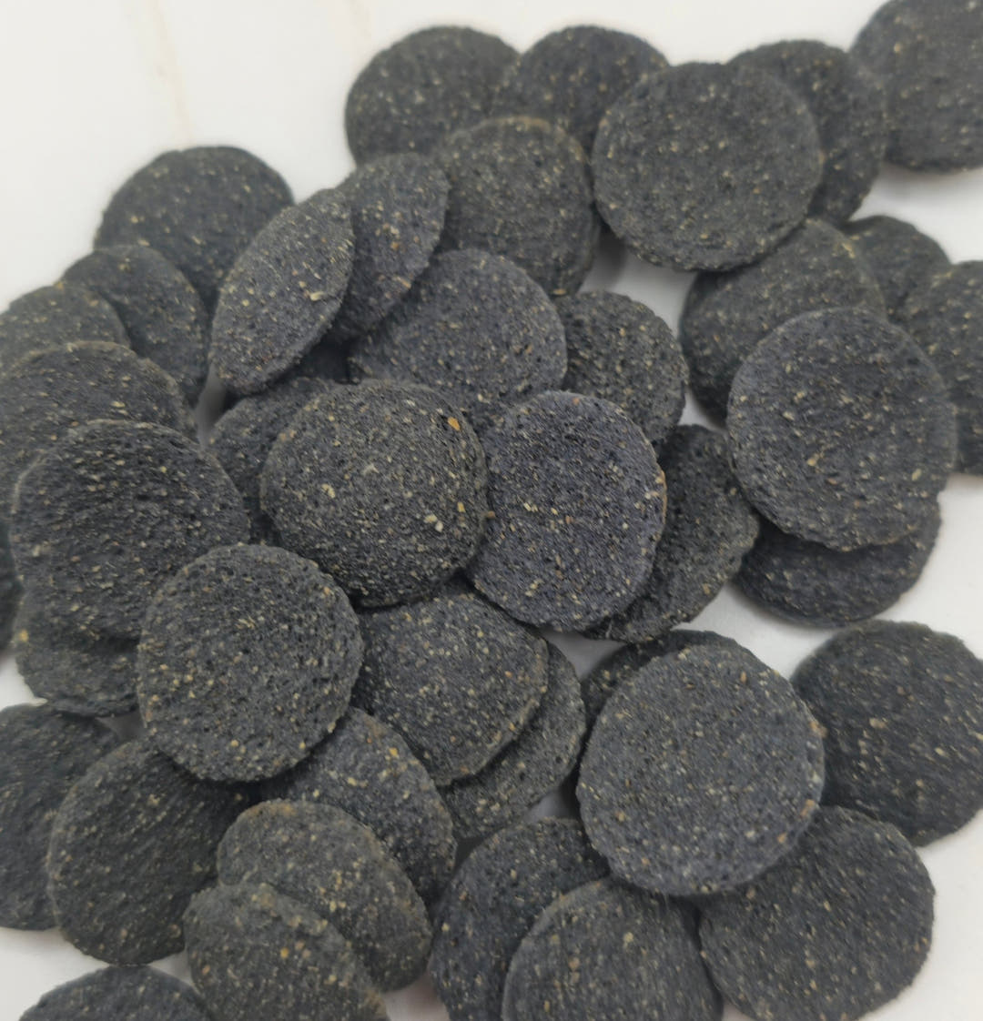 A heap of round Oak and Algae wafers, dark charcoal grey and rough textured, spilling across a white surface.