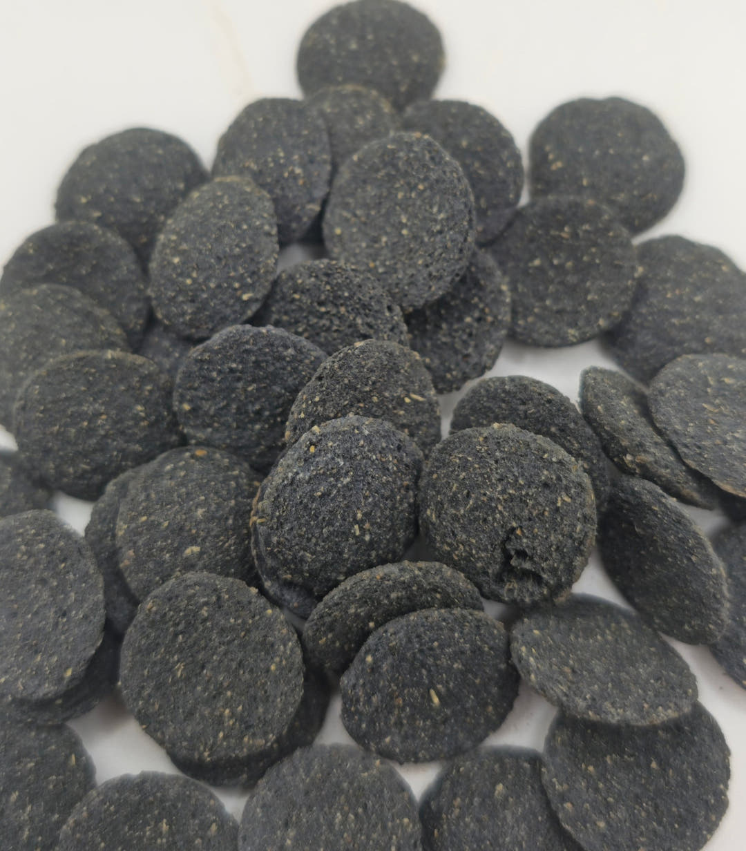 A scattered pile of dark charcoal grey Oak and Algae wafers, each disc speckled with pale flecks, on white.