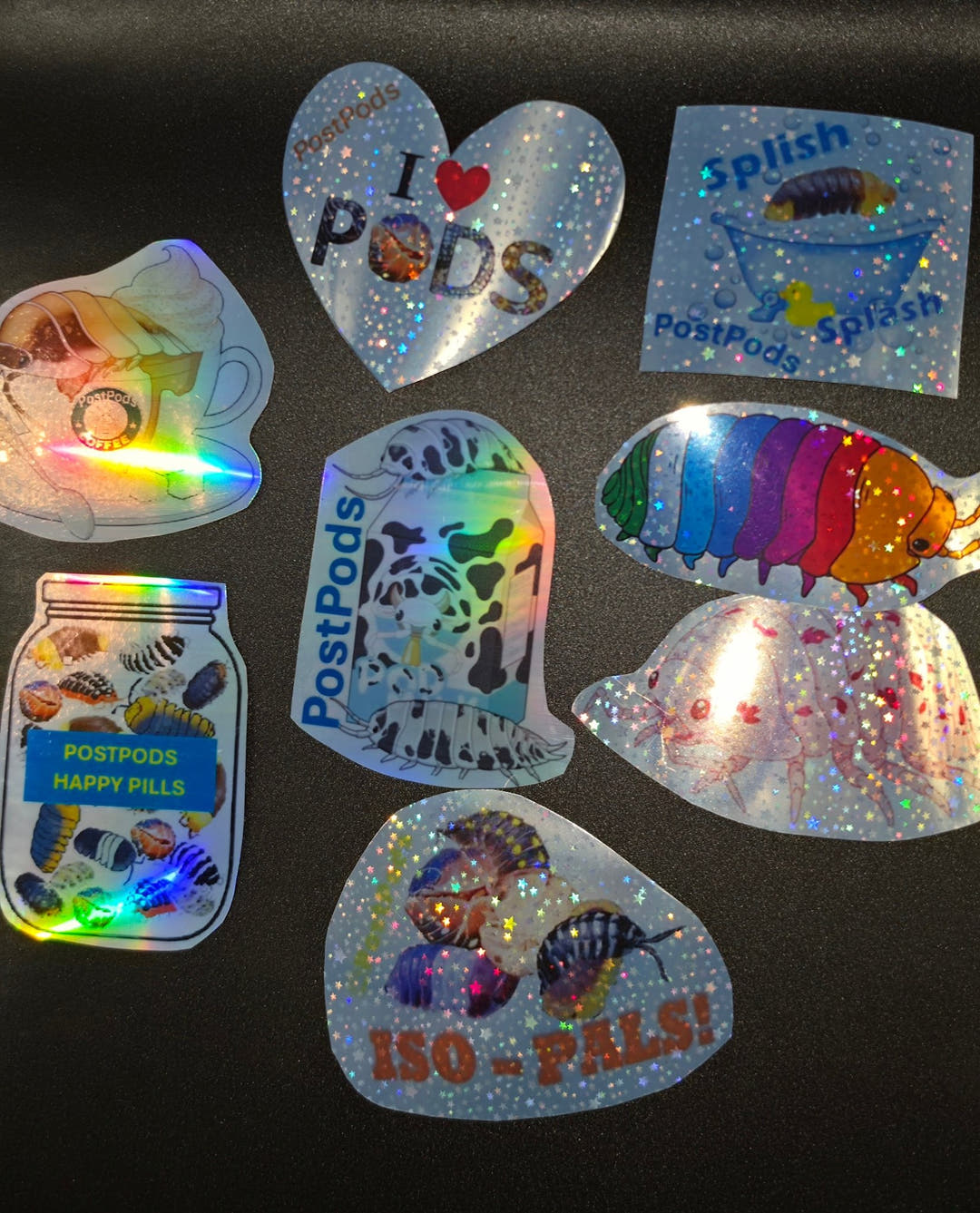 Eight glittery vinyl stickers arranged on a dark surface, among them a heart, a jar and a rainbow isopod.