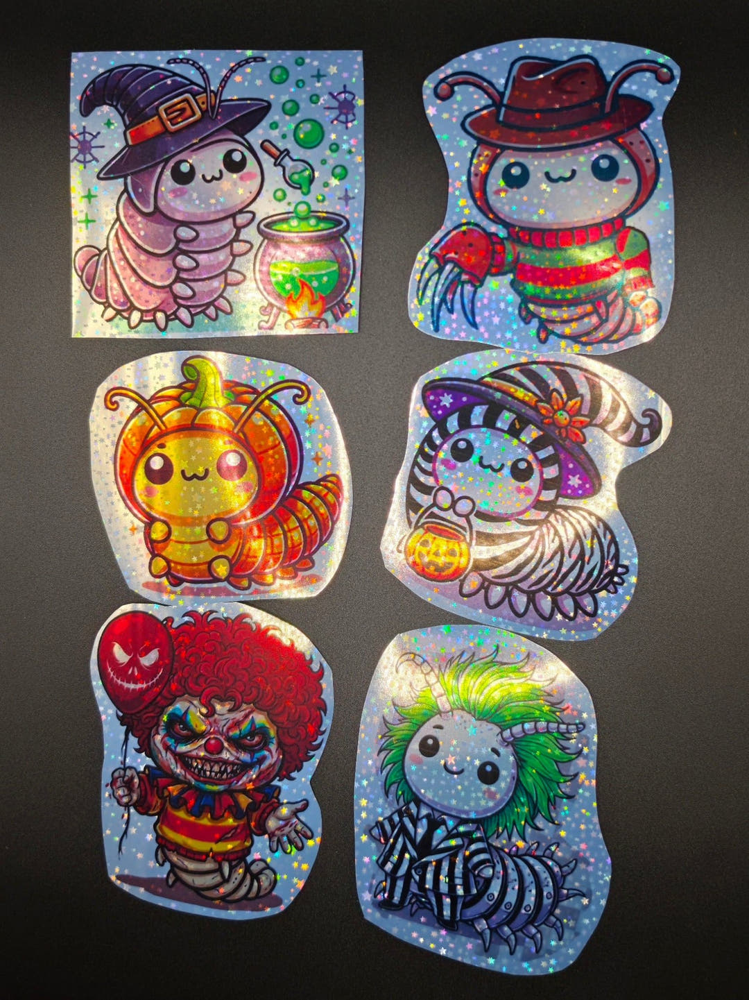 Six glittery Halloween isopod stickers laid out on a dark surface, among them a witch, a pumpkin and a clown.