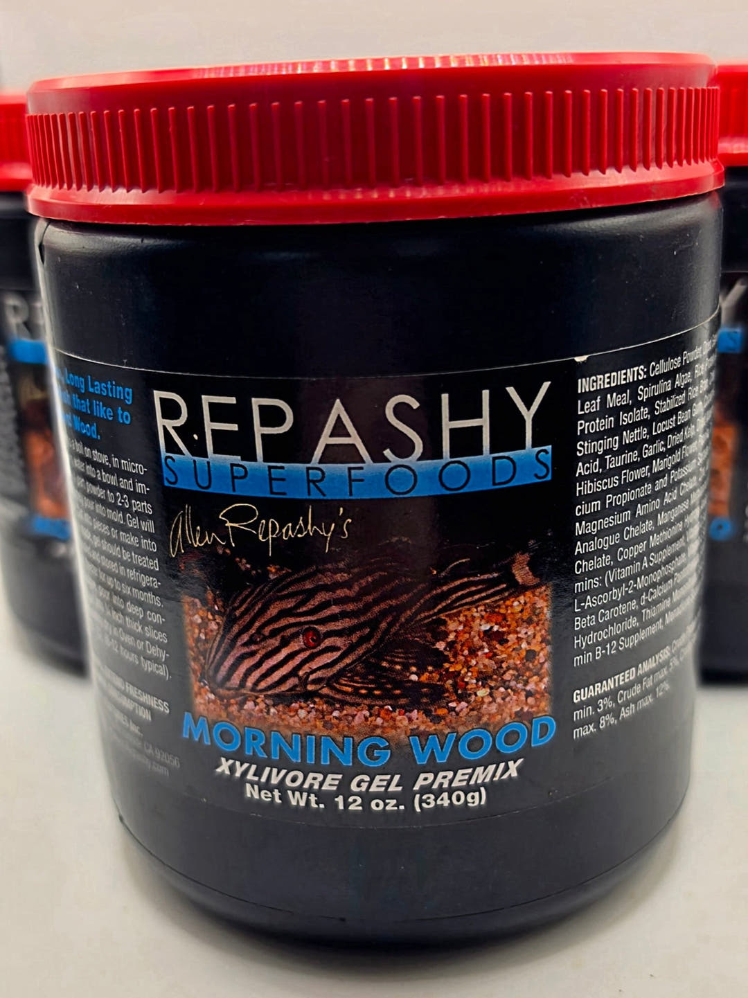 A black Repashy Morning Wood tub with a red lid, front label picturing a striped suckermouth catfish, 12 oz.