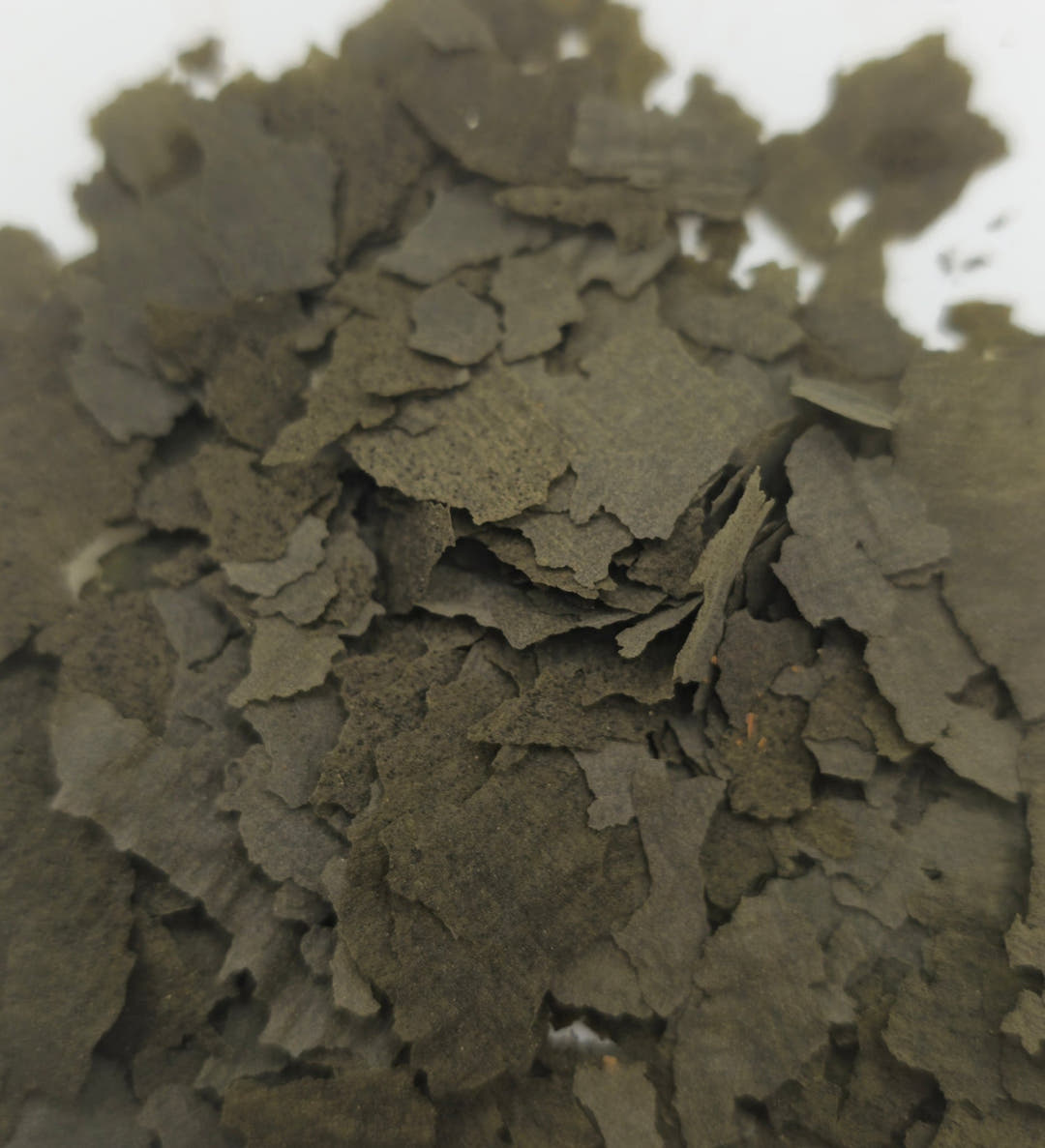 A mound of dark olive spirulina flakes, several pieces standing on edge against white.