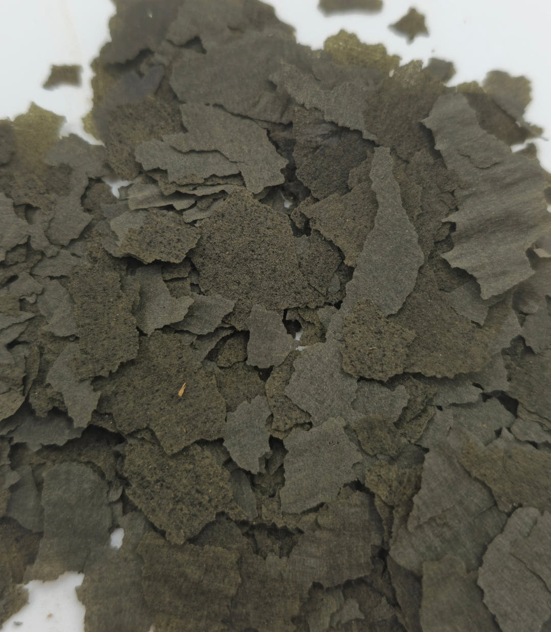 Thin dark green-grey spirulina flakes layered over a white surface, each piece ragged-edged.