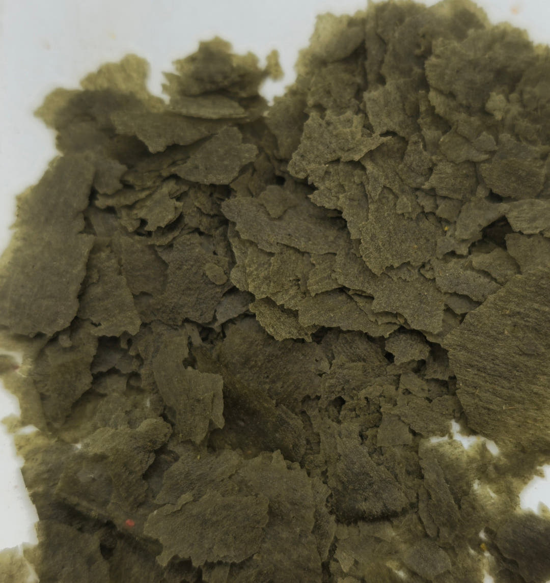 A heap of olive-grey vegetable flakes on a white surface, the pieces overlapping in layers.