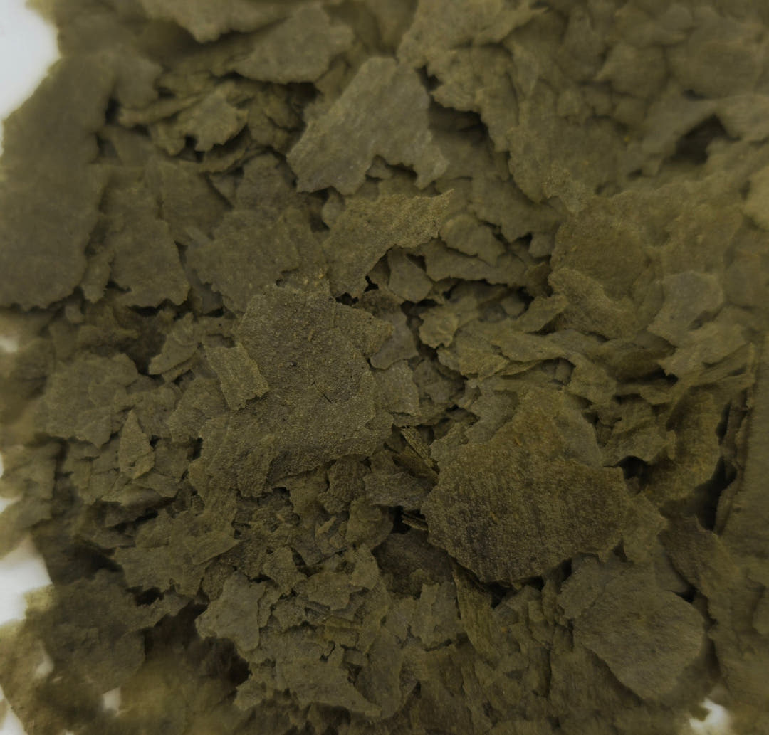 Olive green flakes with spirulina piled loosely, a strip of white surface showing at the left.